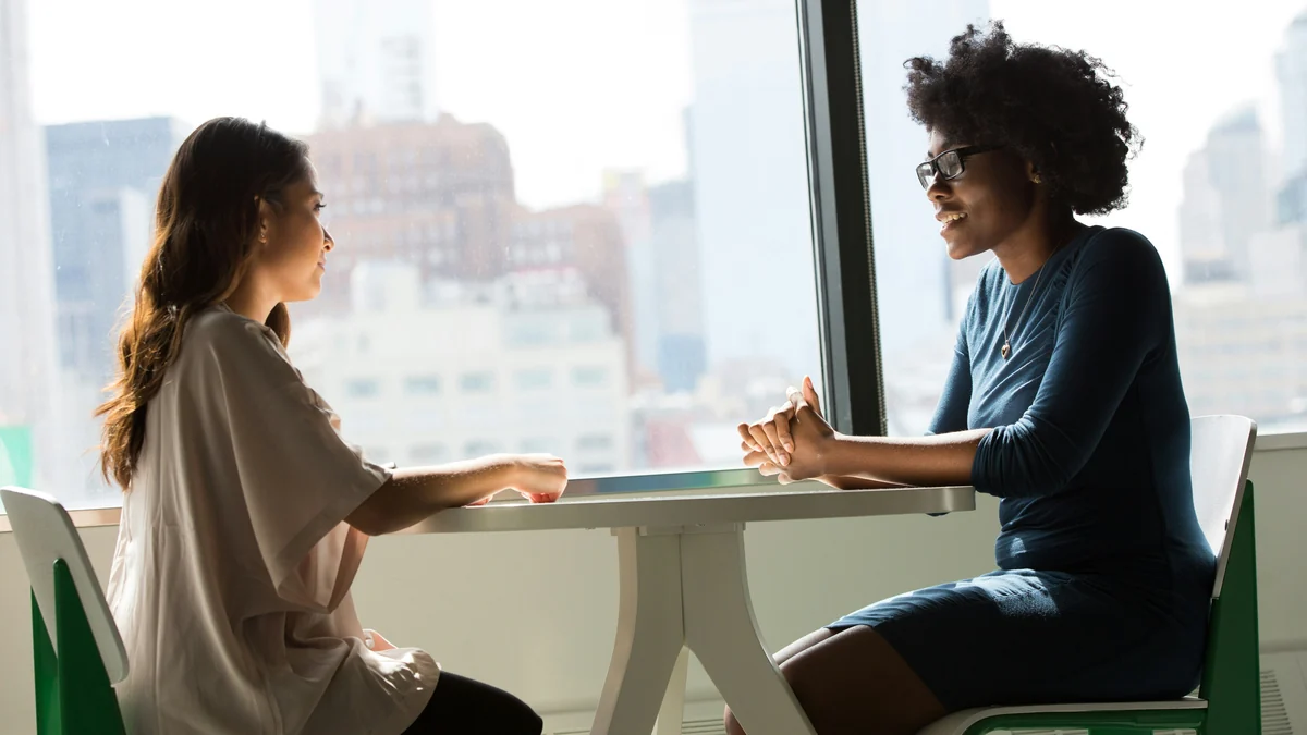 Two women talking face to face at a table beside a window.