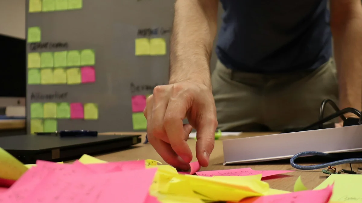 A hand reaching for pink and yellow sticky notes on a table, with grouped notes on a board behind.