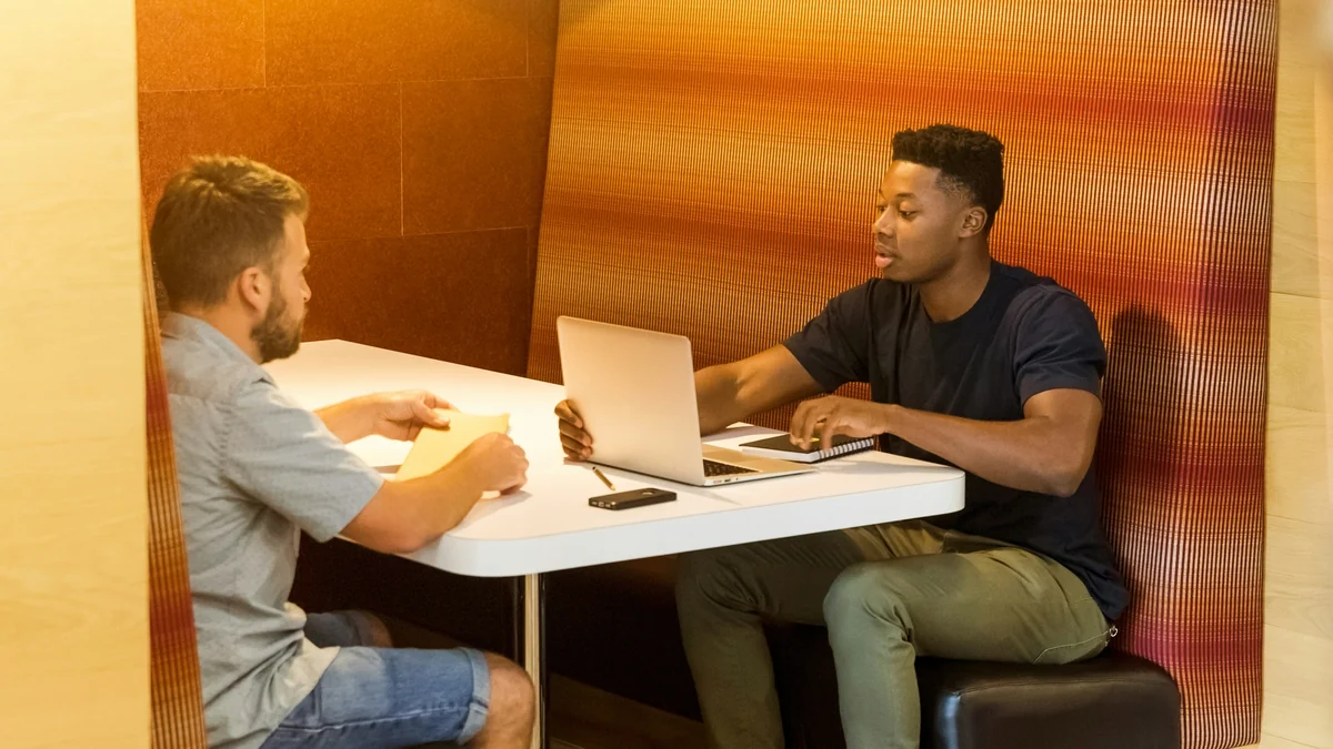 Two colleagues talking across a small table with a laptop and notebook in an office booth.