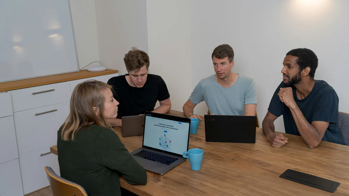 Four colleagues listening and discussing work around a table with open laptops.