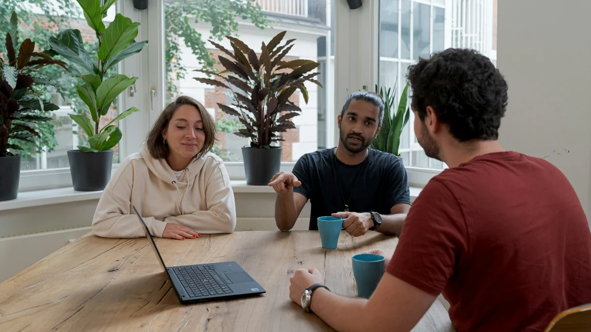 Three colleagues discussing something around a wooden table, with one person gesturing as the others listen.